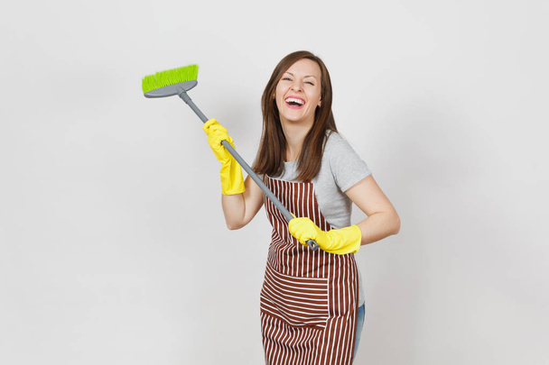 junge lächelnde Hausfrau in gestreifter Schürze, gelbe Handschuhe isoliert auf weißem Hintergrund. Spaß Haushälterin Putzfrau halten und kehren mit Besen. Kopierfläche für Werbefläche - Foto, Bild
