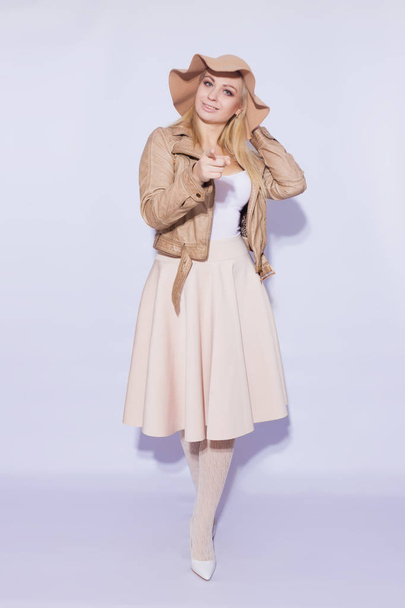 Portrait in full length  beautiful young blond woman. The girl dressed in  midi beige skirt and brown short jacket. On her head is  brown hat. On  legs lace stockings and white shoes with heels. Shows her finger to camera - Photo, image