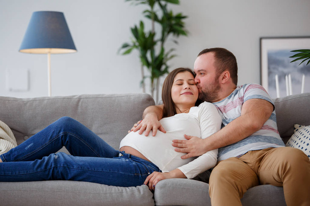 Photo of hugging pregnant woman and man on gray sofa - Фото, изображение