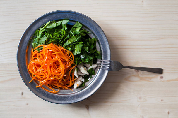 Metallic deep plate filled with spinach, korean carrots and boiled shredded chicken breasts with a small metallic fork  placed on a light natural wooden table surface - Photo, Image