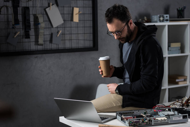 ο άνθρωπος με τον καφέ στο χέρι κάθεται στο τραπέζι και ψάχνετε για φορητό υπολογιστή  - Φωτογραφία, εικόνα