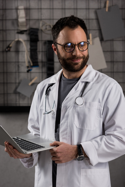 mosolygó férfi orvos fehér kabátot sztetoszkóp át a nyakát, laptop tárolás kezek - Fotó, kép