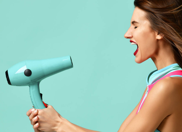 Happy young brunette woman with hair dryer on blue mint background - Valokuva, kuva