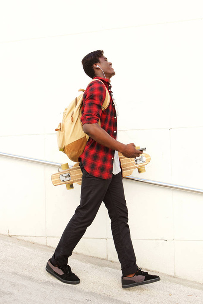 Retrato lateral de cuerpo entero del guapo joven afroamericano estudiante masculino con monopatín y bolso caminando al aire libre
 - Foto, Imagen