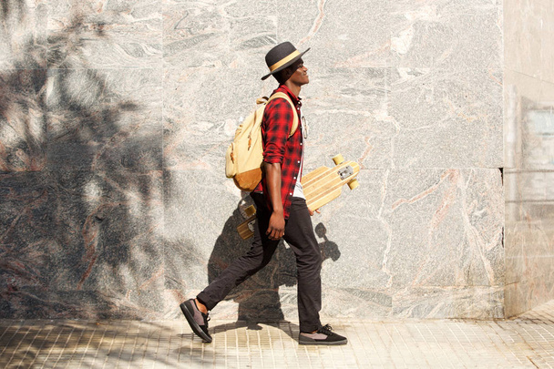 Full length side portrait of young african american man walking outside with skateboard - Фото, зображення