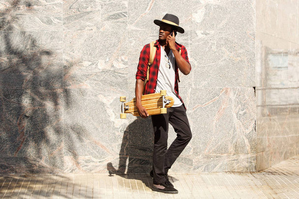 Retrato de comprimento total de belo homem africano com skate falando no celular
 - Foto, Imagem