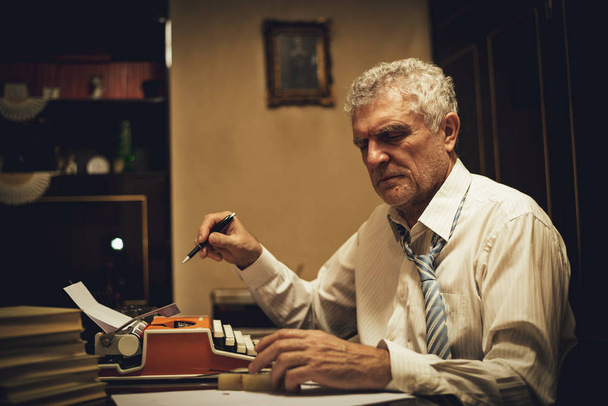 Retro hombre mayor escritor con lápiz en la mano sentado en el escritorio y leyendo un poco de texto para escribir en la máquina de escribir obsoleta
. - Foto, Imagen