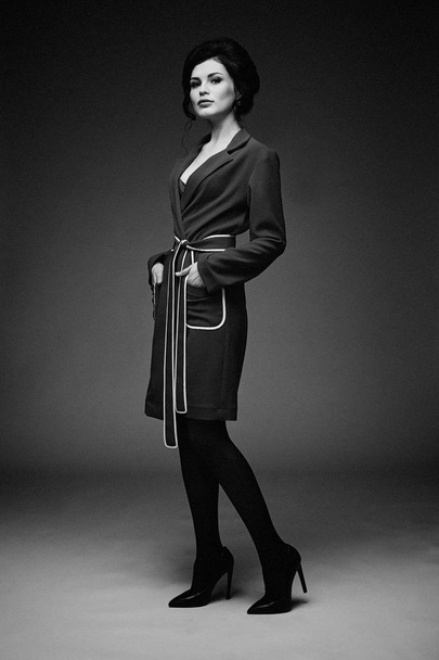 Black and white photo of gorgeous young woman in stylish coat posing against studio background  - Valokuva, kuva