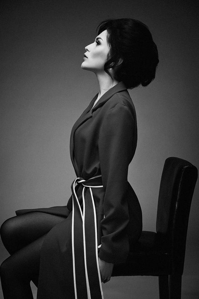 Black and white photo of gorgeous young woman in stylish coat sitting on chair against studio background  - Valokuva, kuva