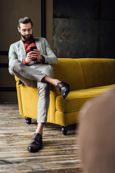 handsome stylish man using smartphone and sitting on yellow couch  - Photo, Image