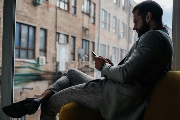 stylish man using smartphone and sitting at window - Zdjęcie, obraz
