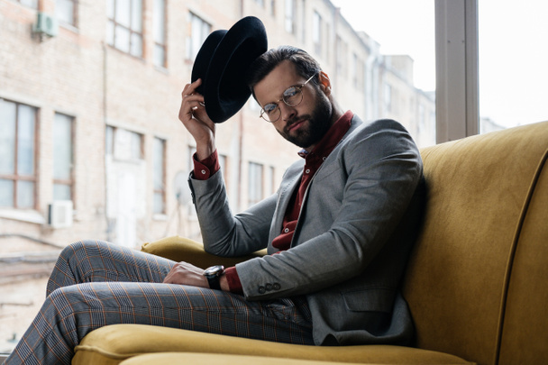 fashionable elegant man in eyeglasses and hat sitting on couch - Fotó, kép