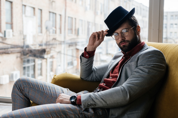 handsome stylish elegant man in eyeglasses and hat sitting on couch - Foto, Imagen