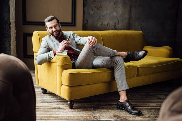 Smějící se elegantní muž s doutníkem a popelník sedí na gauči  - Fotografie, Obrázek