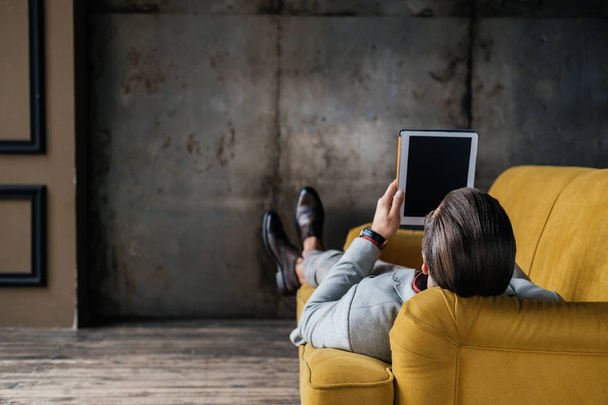 vista posteriore di uomo elegante utilizzando tablet digitale mentre sdraiato sul divano in loft interno
 - Foto, immagini