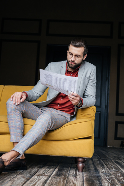 красивий чоловік у модному костюмі читає газету і сидить на дивані
 - Фото, зображення