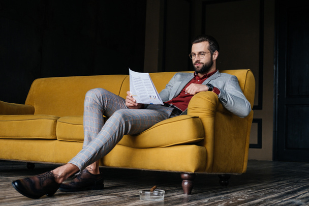 stylish man reading newspaper and sitting on couch, ashtray with cigar on floor - Φωτογραφία, εικόνα