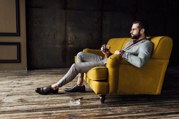 stylish man with glass of whiskey sitting on couch with cigar in ashtray on floor  - Фото, изображение