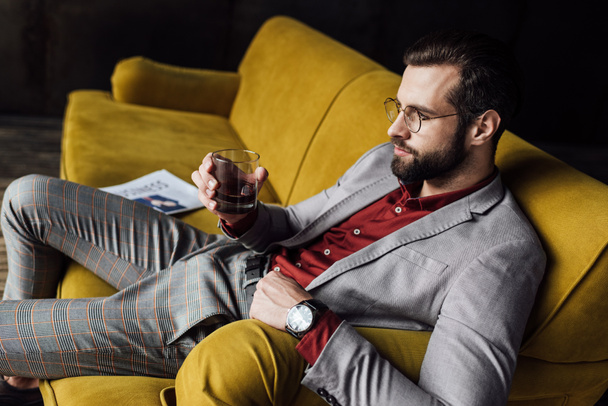 elegant man drinking whiskey and sitting on sofa with newspaper  - Foto, Imagen