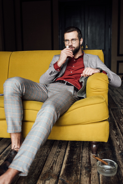 handsome man with glass of whiskey sitting on couch with cigar in ashtray on floor  - Foto, Imagen