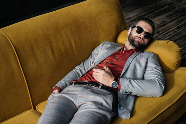 stylish drunk man in sunglasses with glass of cognac lying on yellow sofa - Zdjęcie, obraz