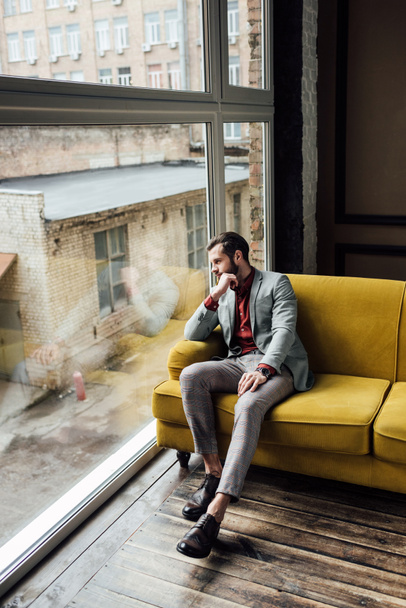stylish thoughtful man sitting on yellow sofa and looking at window  - 写真・画像