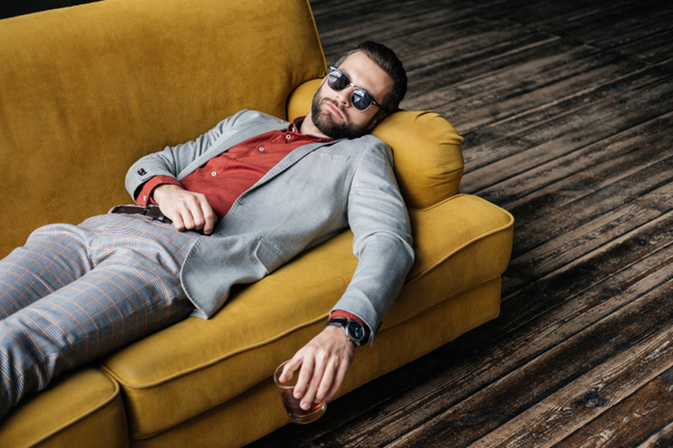 stylish drunk man in sunglasses holding glass of whiskey and lying on yellow sofa - Fotó, kép