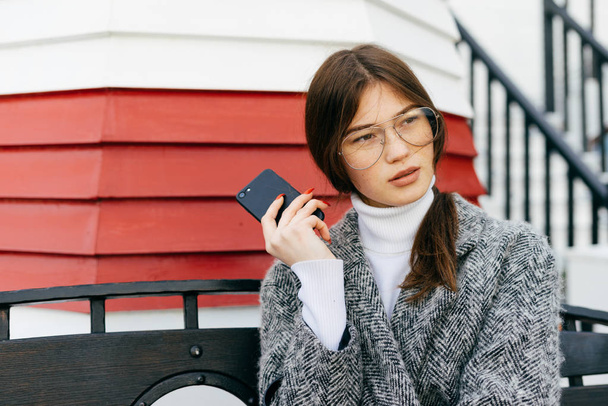 beautiful stylish girl in trendy glasses and a gray coat is sitting on the street, holding a smartphone in her hand - Foto, imagen