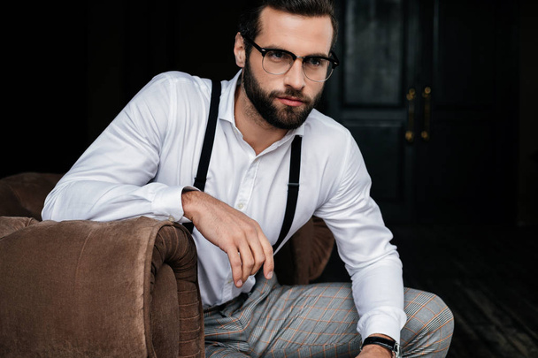 handsome elegant man in eyeglasses, white shirt and suspenders sitting in armchair - Foto, Bild