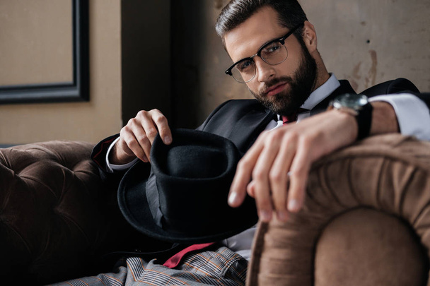 handsome stylish man in eyeglasses holding hat and looking at camera - Photo, Image