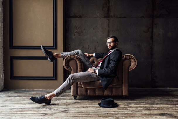όμορφος κομψός επιχειρηματίας θέτοντας σε πολυθρόνα, καπέλο, ξαπλωμένο στο πάτωμα κοντά, εσωτερικό πατάρι  - Φωτογραφία, εικόνα