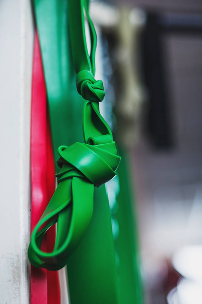rote und grüne Trainingswiderstandsbänder hängen in einer Turnhalle, eines davon in mehreren Knoten gebunden und fokussiert. - Foto, Bild