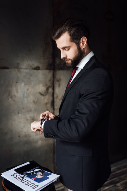 hombre de negocios barbudo mirando el reloj de pulsera mientras está parado en el taburete con el periódico de negocios y el teléfono inteligente
 - Foto, imagen