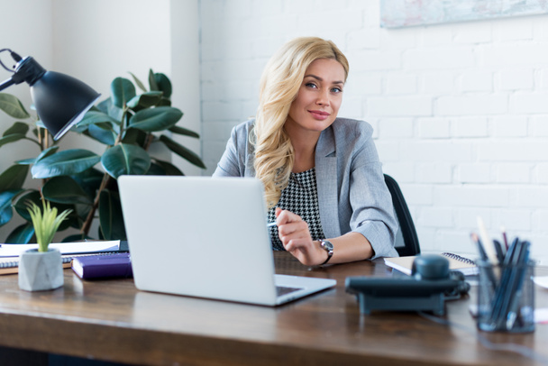 happy businesswoman sitting near laptop and looking at camera - Foto, Bild