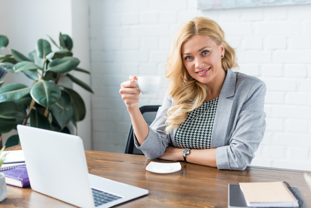 happy businesswoman holding cup of coffee in office and looking at camera - Photo, Image