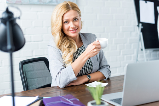 smiling businesswoman holding cup of coffee in office and looking at camera - Valokuva, kuva