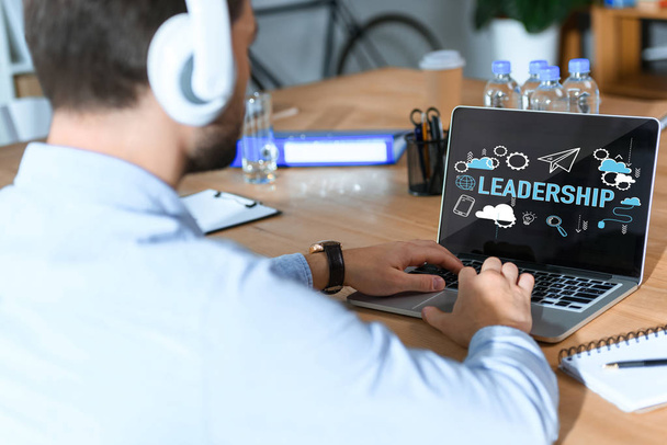 обрезанный снимок бизнесмена в наушниках, слушающего музыку и работающего за ноутбуком со словом лидерства и иконками на экране
  - Фото, изображение