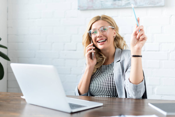 smiling businesswoman talking by smartphone and using laptop in office - Zdjęcie, obraz
