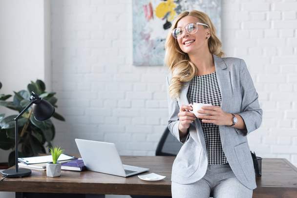 smiling businesswoman holding cup of coffee in office - Photo, image