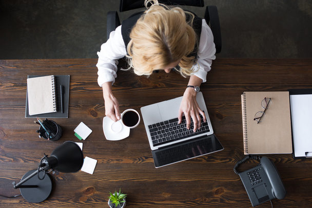vista superior de empresária usando laptop e segurando xícara de café no escritório
 - Foto, Imagem
