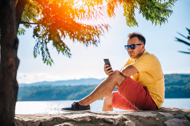 Hombre con camisa amarilla y gafas de sol sentado en el muelle de piedra y el uso de teléfono inteligente
 - Foto, Imagen