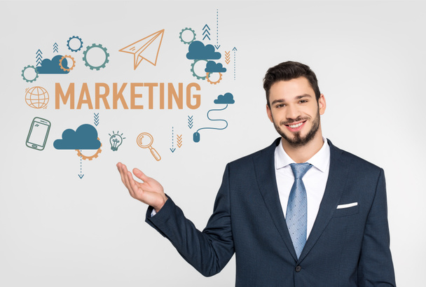 handsome young businessman showing marketing icons and smiling at camera isolated on grey  - Photo, Image