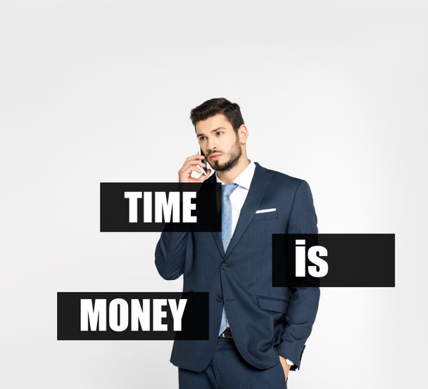 Stilvoller junger Geschäftsmann, der mit der Hand in der Tasche steht und auf dem Smartphone spricht, während er isoliert auf grau blickt, Zeit ist Geld - Foto, Bild