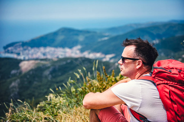 Man with red backpack sitting on top of the mountain and looking at sea horizon - Foto, Imagen