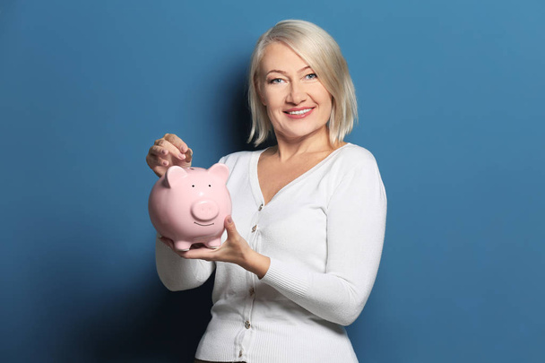 Happy mature woman putting coin into piggy bank on color background - Photo, Image