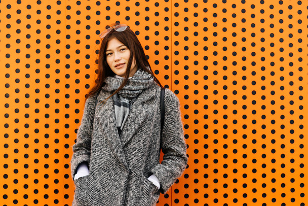 stylish young dark-haired girl in gray coat posing on orange background, street style - Fotografie, Obrázek