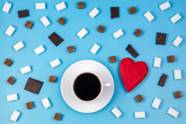 Bílý šálek kávy a červené srdce s kostky hnědé a bílého cukru a čokolády prieces na modrém pozadí - Fotografie, Obrázek