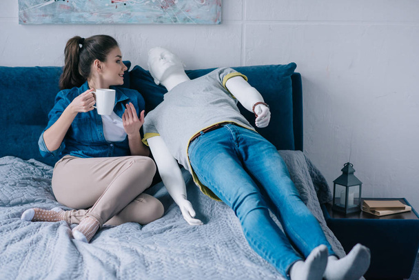 Młoda kobieta z kubkiem kawy rozmowy z manekina podczas odpoczynku na łóżku, koncepcja niespełnionej miłości - Zdjęcie, obraz