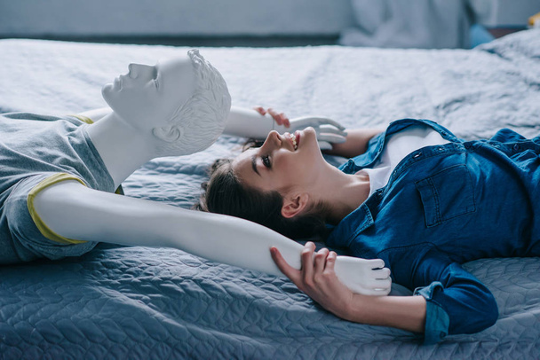 Zijaanzicht van de lachende vrouw en mannequin liggend op bed, eenzaamheid concept - Foto, afbeelding