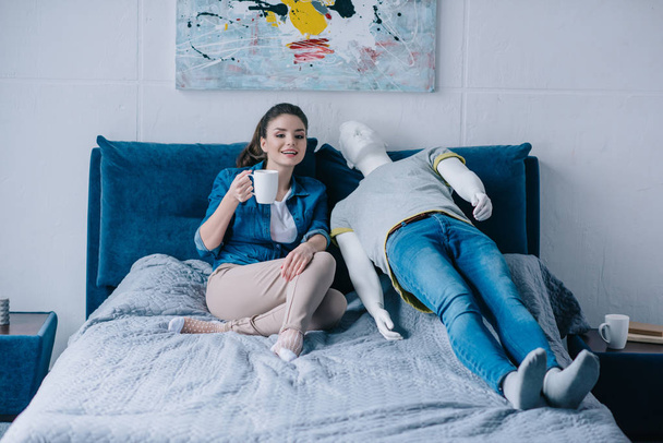 jonge vrouw met kopje koffie praten met etalagepop terwijl rusten op bed, onbeantwoorde liefde concept - Foto, afbeelding
